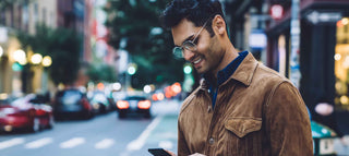 Dark haired man with glasses looks at phone on city street