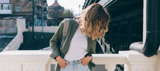 Dirty blonde girl with dry hair in green jacket with hands in pockets of her jeans next to a bridge