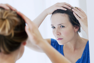 Woman in blue shirt looking closely at hair line in mirror with a white frame