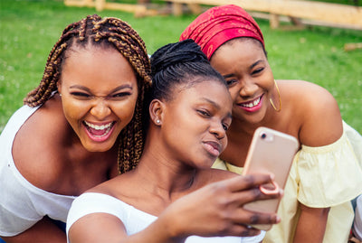 Picture of three beautiful African-American women with box braids, locs, and a turban, taking selfies on a cell phone in the park