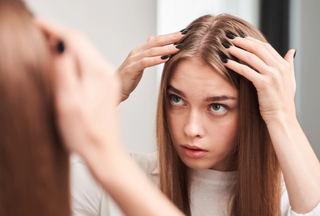 A fair-skinned girl with long strawberry blonde hair wearing a white t-shirt inspects her scalp in a mirror.