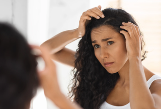 A tan-skinned woman with long curly black hair and dry scalp looking into mirror to check dandruff with distressed look. 
