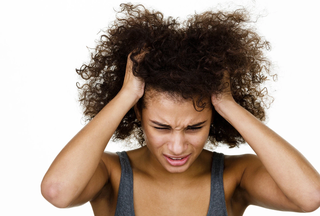 A tan-skinned woman with distressed look has hands in her brown hair itching her scalp in front of a white background.