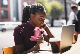 Young African-American woman in maroon top with pulled back braids looks at her laptop at a coffee shop along the street.