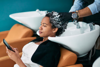  African American woman using smart phone while getting her hair washed at hair salon.
