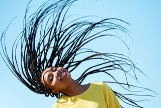 woman smiling wearing a yellow shirt outside with long braided hair and swinging her braids in the air